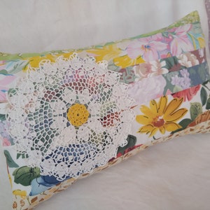 Peut inclure: Coussin décoratif rectangulaire avec un motif patchwork. Le coussin présente des motifs floraux de différentes couleurs, avec un napperon en crochet blanc et un centre jaune. Le coussin mesure environ 40 cm de long.
