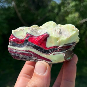 May include: A colorful, layered stone with bands of red, black, and cream colors. The stone has a rough, natural texture and is held in a hand. The stone is a unique decorative item.