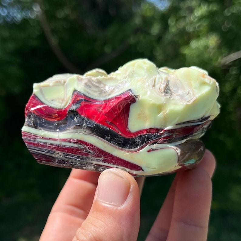 May include: A colorful, layered stone with bands of red, black, and cream colors. The stone has a rough, natural texture and is held in a hand. The stone is a unique decorative item.