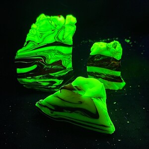 May include: Three irregularly shaped, glowing green objects with dark brown and black marbling. The objects are set against a black background, creating a striking contrast. The shapes appear to be organic, possibly geological specimens.