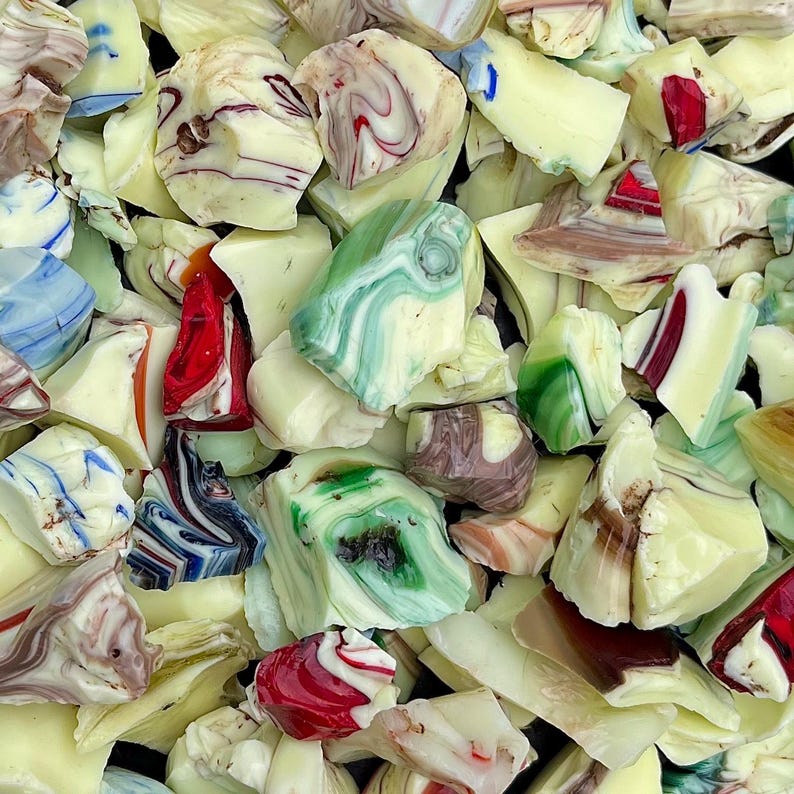 May include: A close-up of a pile of colorful, irregularly shaped glass pieces. The pieces are primarily cream-colored, with swirls of red, green, blue, and brown. The glass has a marbled appearance, with some pieces appearing translucent.