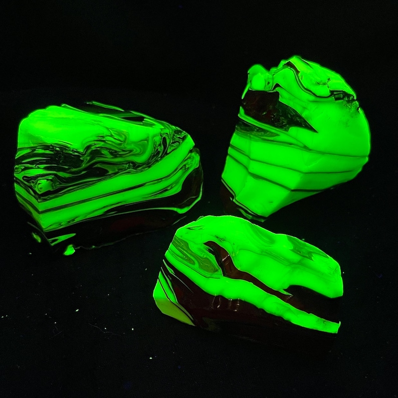 May include: Three glowing green and black marbled rocks against a black background. The rocks have a layered appearance with dark brown and black swirls. The bright green color gives them a luminous quality.