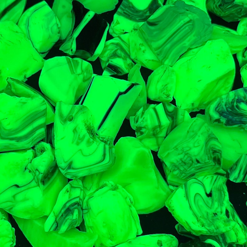 May include: Close-up of a collection of irregularly shaped, glowing green glass rocks. The glass pieces have a marbled appearance with swirls of lighter green and dark green or black. The background is a solid black.