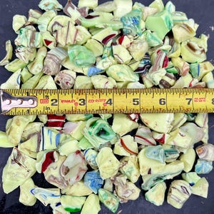 May include: A pile of irregularly shaped, colorful glass pieces with a yellow measuring tape. The glass fragments are primarily pale yellow, with streaks of red, blue, green, and brown. The tape measure shows a length of approximately 6 inches.