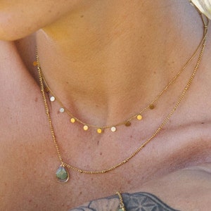 May include: A woman wearing a gold necklace with a teardrop pendant and a delicate chain with small gold circles. She is also wearing a gold ring with a green stone and a gold bracelet with a green stone and a pink stone. Her arms are covered in tattoos.