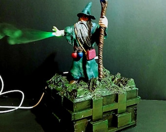May include: A green and brown wizard figurine with a long white beard, holding a staff with a glowing blue tip. The wizard is casting a green spell. The figurine is standing on a green base with a textured surface.