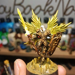 May include: A miniature figurine of a winged warrior with white hair, wearing ornate bronze-coloured armour. The figure holds a glowing yellow sword in one hand and a large shield in the other, standing on a rocky base with a dark circular pedestal.