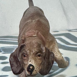 May include: A brown dog figurine with a collar, captured in a forward-facing pose. The dog has floppy ears and a detailed face, with a slightly distressed appearance. The figurine is on a patterned surface.
