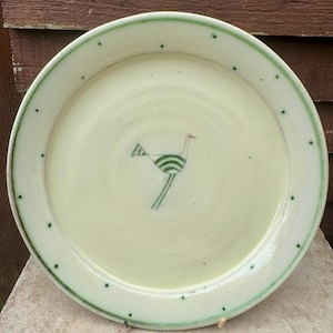 May include: A handmade ceramic plate with a light green background and a green rim. The plate features a whimsical illustration of a bird with green stripes and a red beak. The rim is decorated with small green dots.