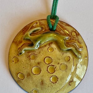 Op de afbeelding: Een ronde keramische hanger met een springende haas, geglazuurd in groen, tegen een gele, maanachtige achtergrond met bruine accenten. De hanger heeft een getextureerd oppervlak en hangt aan een groen koord. Rustiek ontwerp.