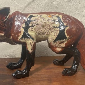 May include: A ceramic fox ornament with a glossy finish. The fox is mainly brown with black legs and a white-tipped tail. The fox's body features a detailed landscape design with trees and a sun. The fox is posed in a walking stance.