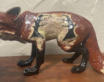 Op de afbeelding: Een keramieken vosfiguur met een glanzende afwerking. De vos is voornamelijk bruin met zwarte poten en een witgepunte staart. Het lichaam van de vos heeft een gedetailleerd landschapsontwerp met bomen en een zon. De vos staat in een loopstand.