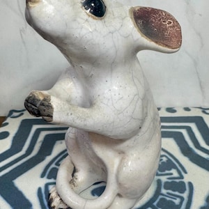 May include: A ceramic mouse figurine in a seated pose. The mouse is mainly white with dark eyes, brown ears, and dark accents on its paws and tail. The figurine has a cracked glaze finish, giving it a vintage appearance.