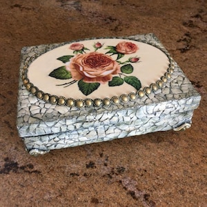 May include: A decorative wooden box with a mosaic-like finish. The lid features a floral design with a large pink rose and smaller pink roses. The box has a gold trim and decorative feet.
