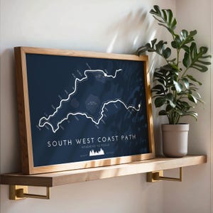 May include: A framed print depicting the South West Coast Path from Minehead to Poole, with a navy blue backdrop and a white path. The print is in a light wood frame, on a wooden shelf with brass brackets. A potted plant is to the right.
