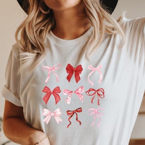 Op de afbeelding: Wit T-shirt met een afbeelding van roze en rode strikken. De strikken zijn in verschillende stijlen en maten.