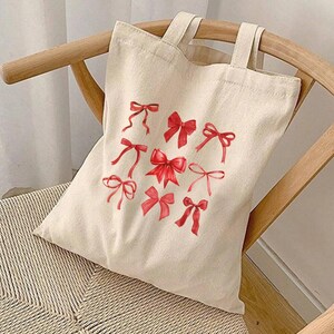 Op de afbeelding: Een witte canvas tote bag met rode strikken erop gedrukt. De tas heeft twee handvatten en staat op een houten stoel.