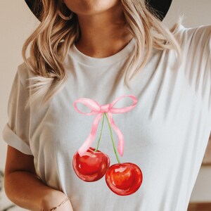 May include: White t-shirt featuring a pink ribbon and two red cherries. The cherries have green stems and are tied together with a pink ribbon.