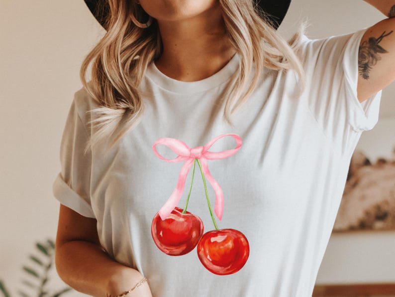 May include: White t-shirt featuring a pink ribbon and two red cherries. The cherries have green stems and are tied together with a pink ribbon.