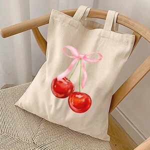 May include: A beige canvas tote bag featuring a pink ribbon and two red cherries printed on it.