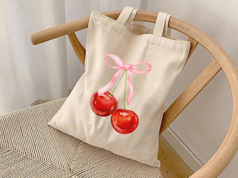 May include: A beige canvas tote bag featuring a pink ribbon and two red cherries printed on it.