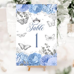 May include: A table number card with a floral and butterfly design. The card features blue flowers, silver butterflies, and the word "Table" with the number "1" in a blue font. The card is displayed on a small wooden easel.