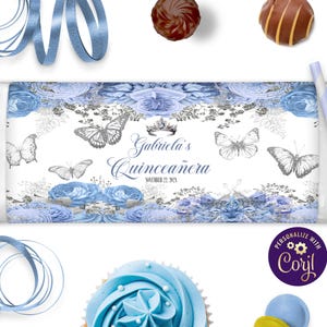 May include: A white candy bar wrapper with a floral design in shades of blue and silver, featuring butterflies and the text "Gabriela's Quinceañera". Includes a crown graphic and the date November 22, 2025. Decorative elements include blue ribbons, a cupcake, and candies.