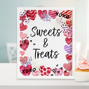 May include: White framed sign with the text "Sweets & Treats" surrounded by a border of pink, red, and purple watercolor hearts. The sign is sitting on a white surface next to a pink frosted cupcake.