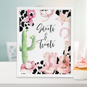 May include: A printable sign with a pink and black cow print background. The sign says "Sweets & Treats" in black script. There are pink cowboy boots, a pink guitar, a pink horseshoe, a pink star, and a cactus on the sign.