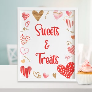 May include: White framed sign with red hearts and gold accents. The sign says "Sweets & Treats". A pink frosted cupcake with sprinkles sits on a white surface in front of the sign.