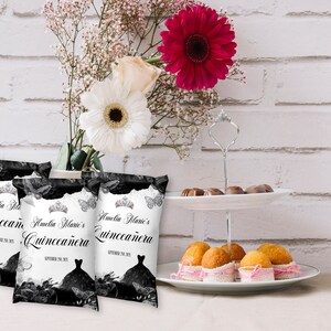 May include: Three black and white favour bags with the text "Amelia Marie's Quincea&ntilde;era" and a black silhouette of a dress with a crown on top. The bags are on a white surface with a white brick wall in the background.