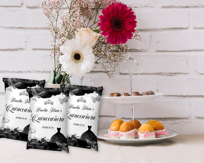 May include: Three black and white favour bags with the text "Amelia Marie's Quincea&ntilde;era" and a black silhouette of a dress with a crown on top. The bags are on a white surface with a white brick wall in the background.