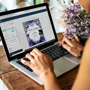 May include: A laptop displaying a digital invitation design with a dark blue and black color scheme. The invitation features floral and spiderweb graphics, with the names "Martha & Gregory" and event details. The laptop is on a wooden table.
