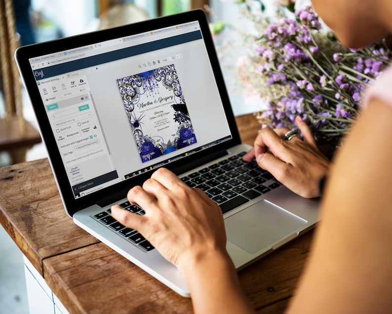 May include: A laptop displaying a digital invitation design with a dark blue and black color scheme. The invitation features floral and spiderweb graphics, with the names "Martha & Gregory" and event details. The laptop is on a wooden table.
