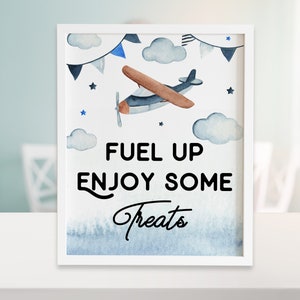 May include: A white framed sign with a blue and gray watercolor background featuring a cartoon airplane flying through clouds. The sign says "Fuel Up Enjoy Some Treats" in black text.