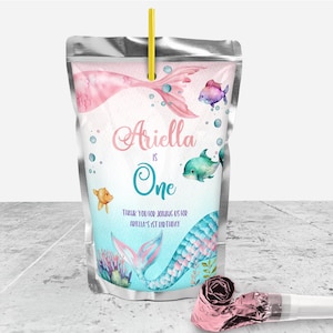 May include: A silver juice pouch with a mermaid tail design. The pouch has a pink and blue mermaid tail with a yellow straw sticking out of the top. The pouch has the text "Ariella is One" and "Thank you for joining us for Ariella's 1st birthday".