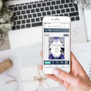 May include: A smartphone displays a wedding invitation design with a blue and black floral theme. The invitation includes the names "Martha & Gregory" and event details. The phone is held over a white surface with a laptop and flowers.