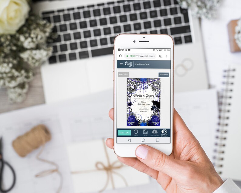 May include: A smartphone displays a wedding invitation design with a blue and black floral theme. The invitation includes the names "Martha & Gregory" and event details. The phone is held over a white surface with a laptop and flowers.