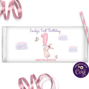 Può includere: Confezione di barretta di cioccolato bianco con un coniglietto rosa che tiene un palloncino rosa numero uno. La confezione ha il testo "Emily's First Birthday" e "Thanks for Hopping By!"