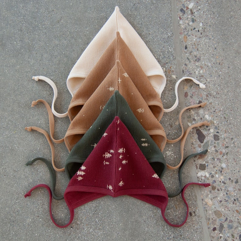 May include: Five waffle-knit baby bonnets in a stacked arrangement. The bonnets are in shades of cream, tan, olive green, and burgundy. Each bonnet has a pointed top and ties. The burgundy bonnet has a white tree pattern.