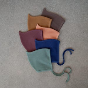 May include: A collection of six waffle-knit baby bonnets in various colors, including teal, navy, peach, mustard, plum, and brown. Each bonnet has a rounded shape and ties. The bonnets are arranged in a fan shape on a gray surface.
