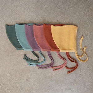 May include: A collection of six ribbed knit bonnets arranged in a row, featuring a spectrum of colors including dark green, light green, mauve, dusty rose, terracotta, and mustard yellow, each with matching fabric ties at the base.