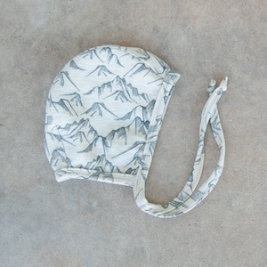 May include: A light-colored baby bonnet with a mountain range pattern in gray. The bonnet has ties on each side and is made of soft fabric. The mountain design covers the entire bonnet.