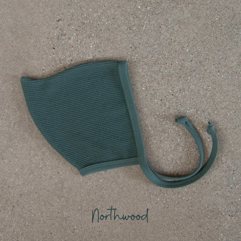 May include: A teal-colored baby bonnet with a textured waffle knit design. The bonnet has a rounded shape with a brim and ties. The word "Northwood" is written in a script font at the bottom of the image.