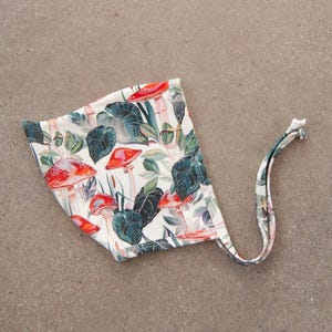 May include: A white baby bonnet with a colorful mushroom and leaf pattern. The bonnet features red mushrooms, green leaves, and a tie closure. The fabric has a botanical design.