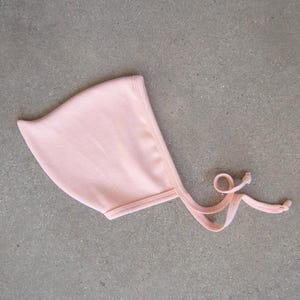 May include: A pale pink baby bonnet made from ribbed knit material. The bonnet features a rounded design with a small brim and ties for fastening under the chin. The ties are finished with knots. It rests on a grey surface.