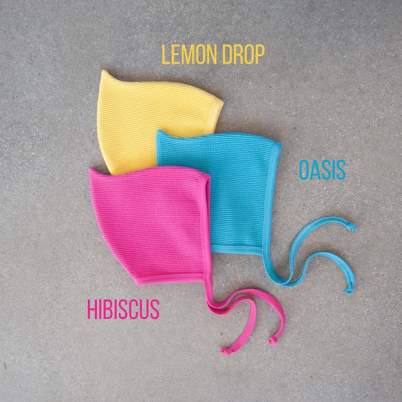 May include: Three colorful bonnets in yellow, turquoise, and pink, with the text "Lemon Drop", "Oasis", and "Hibiscus". Each bonnet has a textured knit fabric and ties. The bonnets are displayed on a gray surface.