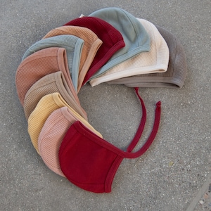 Peut inclure: Une collection de bonnets pour bébés de différentes couleurs, dont le rouge, le jaune et le bleu. Les bonnets sont faits d'un tissu texturé et ont des liens sous le menton. Les bonnets sont disposés en cercle.