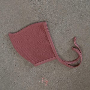 May include: A fig-colored baby bonnet with a waffle knit texture. The bonnet has a rounded shape with a stitched edge and long, thin ties. The word "Fig" is written in cursive at the bottom.