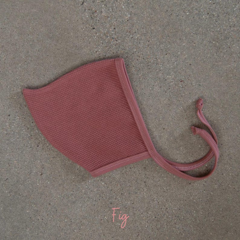 May include: A fig-colored baby bonnet with a waffle knit texture. The bonnet has a rounded shape with a stitched edge and long, thin ties. The word "Fig" is written in cursive at the bottom.